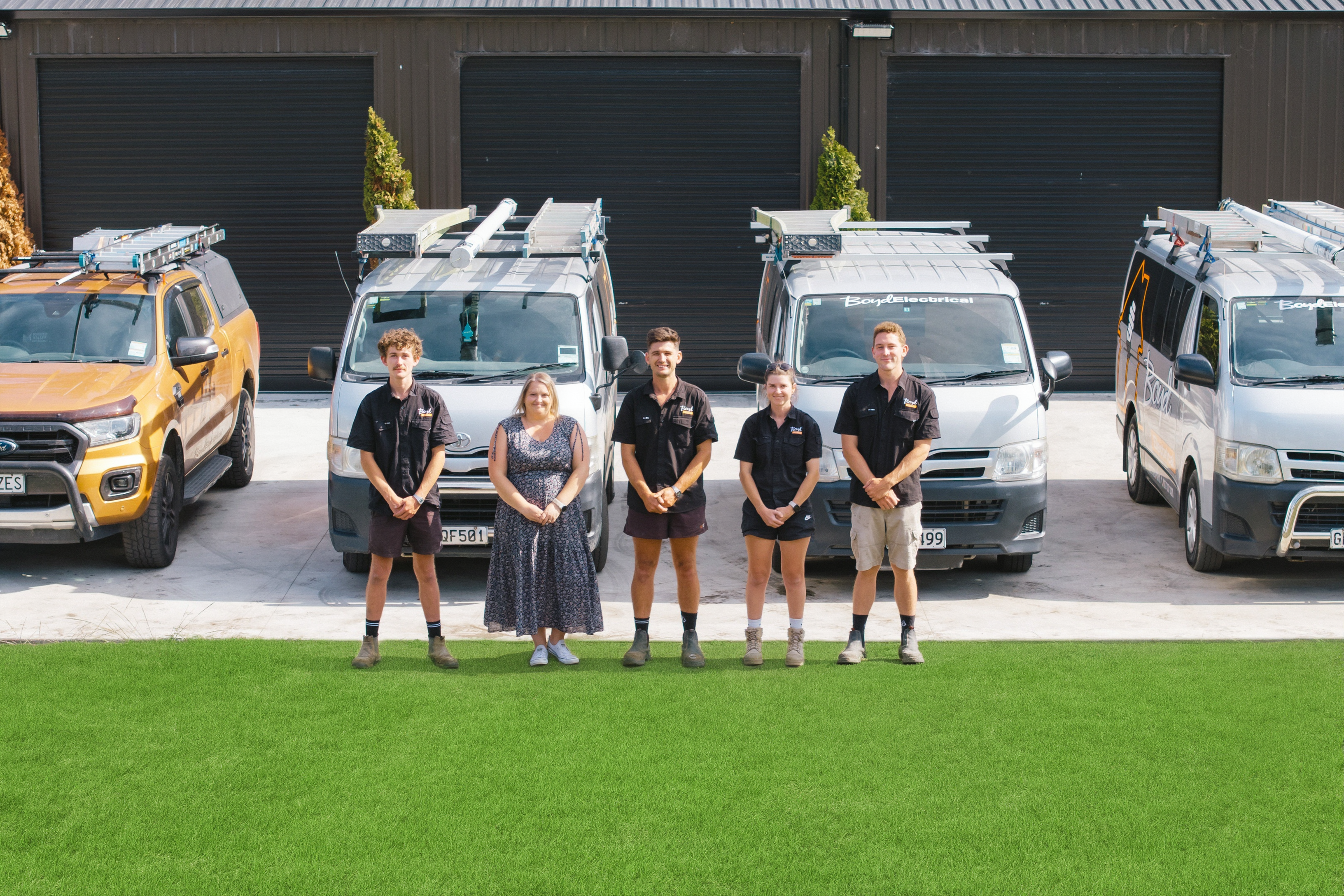  Boyd Electrical Whanganui team of five standing in front of their fleet of branded work vehicles and workshop